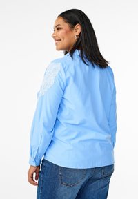 Blusa azzurro chiaro con maniche lunghe, caratterizzata da dettagli in pizzo sulla spalla sinistra. Design semplice, texture liscia, abbinata a jeans.