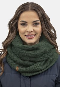Bufanda de infinito de punto verde oscuro con un patrón texturizado y una pequeña etiqueta color beige, llevada sobre una chaqueta azul marino.