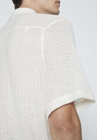 Camisa blanca de manga corta con textura tejido y corte holgado, que presenta collar y costuras visibles en las mangas y la espalda.