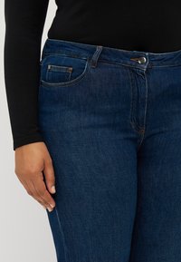 Jean en denim bleu foncé avec une coupe classique, un design à cinq poches et des coutures contrastantes orange le long des coutures. La ceinture est dotée d'un bouton en métal.