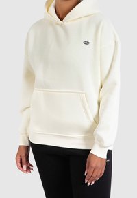 Cremefarbener Hoodie aus weichem Stoff, mit einer Fronttasche, gerippten Bündchen und einem kleinen Logodetail auf der Brust.