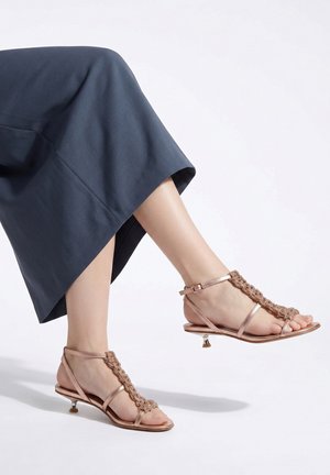 Piedi che indossano sandali rosa oro con cinturini, piccoli tacchi e dettagli intrecciati, abbinati a una gonna blu scuro su sfondo bianco.