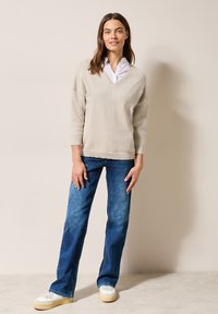 Helles beige V-Ausschnitt-Pullover mit abfallenden Schultern, kombiniert mit blauen gerade geschnittenen Jeans und weißen Sneakers. Einfaches Design und lässige Passform.