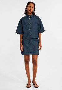 Denimoutfit met een korte mouw en een kraagtop en een bijpassende kokerrok. Beide zijn donkerblauw met verticale strepen en knoopdetails.