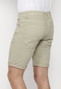 Homme portant un short beige retroussé et une chemise blanche, vu de dos au niveau des cuisses sur un fond neutre.