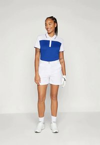 Modra in bela golf polo s gumbno zaponko, v kombinaciji z belimi kratkimi hlačami. Gladek material, kratke rokave in minimalni dodatki.