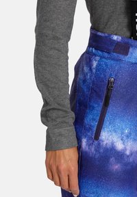 Broek met een melkwegpatroon in een blauw en paars kleurenpalet, voorzien van een ritszak en een klittenbandsluiting. Grijze lange mouw top zichtbaar.