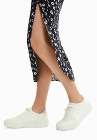 Baskets en cuir blanc avec des surfaces texturées, bout rond et design à lacets. Portées avec une jupe noire à motifs floraux avec une fente latérale.