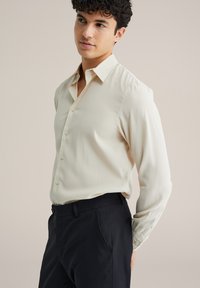 Svetlo béžová košeľa na gombíky s klasickým golierom a dlhými rukávmi, vyrobená z hladkej látky, v kombinácii s tmavými nohavicami.