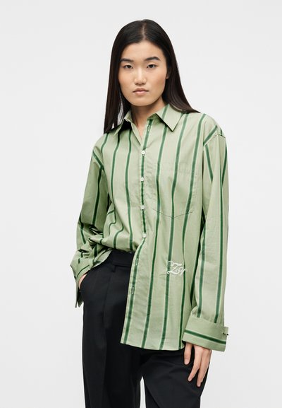 Νεαρή γυναίκα με μακριά σκούρα μαλλιά, φορώντας ένα άνετο ανοιχτό πράσινο πουκαμισάκι με ρίγες και μαύρο παντελόνι, στέκεται με το ένα χέρι στην τσέπη.