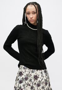 Μαύρο μακρυμάνικο τοπ με ψηλό γιακά, με υφή υφάσματος. Συνδυάζεται με φούστα με λουλουδάτο μοτίβο σε λευκούς, μωβ και πράσινους τόνους.