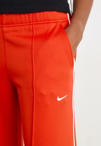 Røde treningbukser laget av glatt stoff, med hvite sidestriper og en liten hvit Nike-logo på venstre lår.