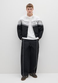 Zwart-wit kleurgeblokte jas met ritssluiting en elastische manchetten, gecombineerd met losse zwarte cargobroek met witte biezen en zakken.