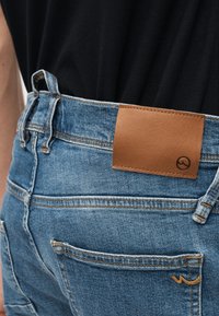 Άτομο που φορεί μπλε τζιν με ταμπά δερμάτινο ένθετο και μαύρο πουκάμισο μαζεμένο στη μέση.