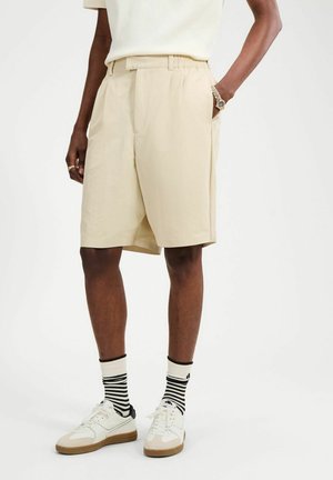 Persoon draagt beige knielange shorts, witte sneakers, zwart-wit gestreepte sokken, een wit overhemd en een gouden polshorloge.