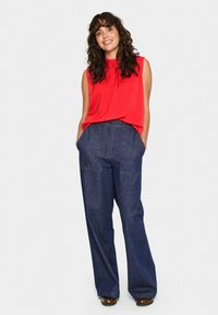 Blusa rossa senza maniche con un dettaglio a fiocco sul collo, abbinata a pantaloni in denim blu a vita alta e gamba larga, con tasche frontali.