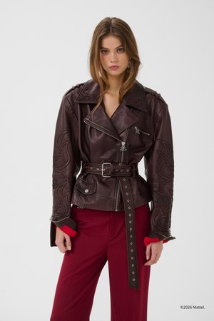 Mladá žena v tmavohnedej koženej bunde s opaskom, zdobenej reliéfnymi vzormi, a červených širokých nohaviciach, stojaca pred jednofarebným pozadím.