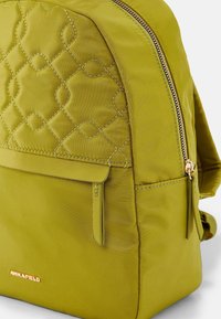 Olive green backpack with quilted pattern on front pocket, gold zipper, and "ANNA FIELD" logo at the bottom.