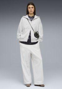 Γυναίκα στέκεται προς τα εμπρός, φορώντας ανοιχτό γκρι φούτερ με κουκούλα, φαρδιά λευκά παντελόνια, μαύρο πουκάμισο, μπεζ παπούτσια και μικρή μαύρη τσάντα χιαστί με λογότυπο.