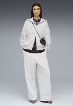 Γυναίκα στέκεται προς τα εμπρός, φορώντας ανοιχτό γκρι φούτερ με κουκούλα, φαρδιά λευκά παντελόνια, μαύρο πουκάμισο, μπεζ παπούτσια και μικρή μαύρη τσάντα χιαστί με λογότυπο.