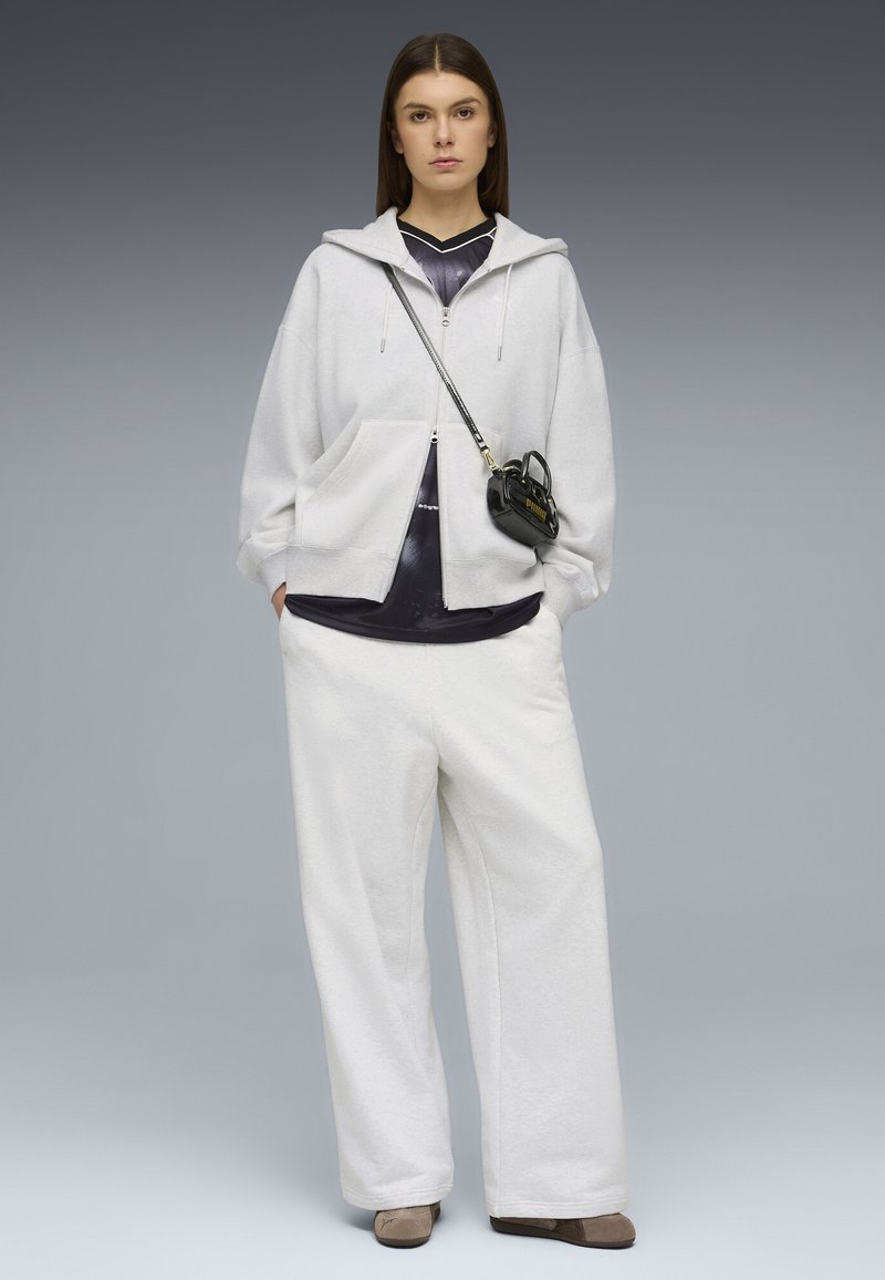 Γυναίκα στέκεται προς τα εμπρός, φορώντας ανοιχτό γκρι φούτερ με κουκούλα, φαρδιά λευκά παντελόνια, μαύρο πουκάμισο, μπεζ παπούτσια και μικρή μαύρη τσάντα χιαστί με λογότυπο.