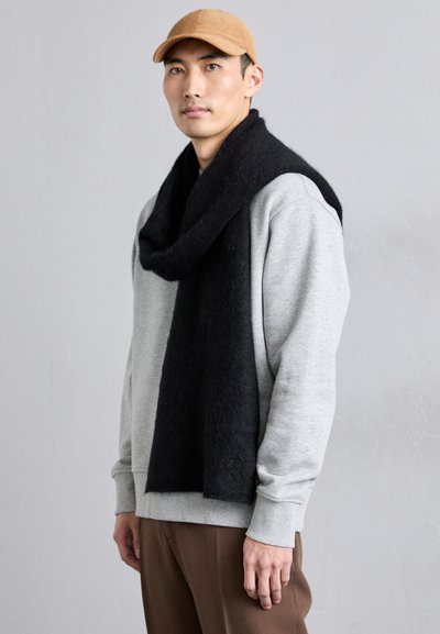 Sudadera de algodón gris con hombros caídos, bufanda de lana negra caída alrededor del cuello y gorra color beige. Textura suave, corte casual.