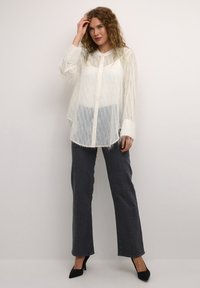 White sheer shirt with textured fringes, button front, paired with dark denim pants featuring silver accents and black heels.