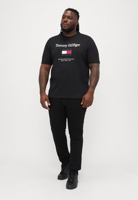 Must mustärgne lühikese varrukaga T-särk, millel on valge "Tommy Hilfiger" bränding ja väike logo trükk punases, valges ja sinises. Ühendatud mustade teksade ja spordijalatsitega.