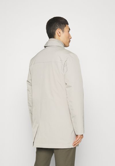 Chaqueta impermeable de color gris claro, con cuello alto, aberturas laterales y diseño minimalista. Presenta una textura suave y un sutil logotipo en la parte de atrás.