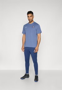 Blå atletisk t-shirt med logo og korte ærmer, parret med blå atletiske leggings og sorte fodboldstøvler. Glat tekstur, tætsiddende design.