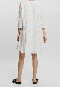 Vit bomullsklänning med lagerdesign, ögonlet-detaljer och klockärmar. Bärs med svarta platta sandaler och visar en avslappnad passform.