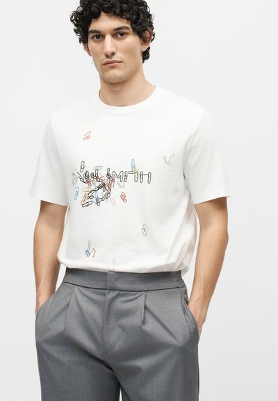 Νέος άνδρας με σγουρά μαλλιά που φοράει λευκό μπλουζάκι με πολύχρωμο μοτίβο συνδετήρων και γκρι πλισέ παντελόνια, με τα χέρια στις τσέπες.