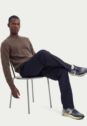 Uomo che indossa un maglione marrone, pantaloni blu navy e scarpe da ginnastica, seduto a gambe incrociate su una sedia di metallo davanti a uno sfondo semplice.