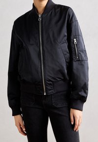 Černá bomber bunda vyrobená z hladkého, lehkého materiálu. Má přední zip, žebrované manžety a lem, a zipovou kapsu na rukávu.