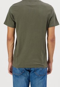 Bavlněné tričko olivově zelené barvy s krátkými rukávy, kulatým výstřihem a přímým lemem, nošené s modrými džínovými kalhotami, pohled zezadu.