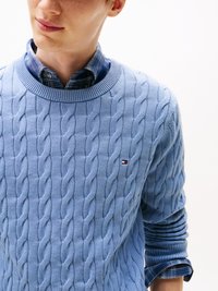 Suéter azul-claro em malha trançada, com decote redondo canelado e padrão texturizado. Combinado com uma camisa xadrez azul por baixo.