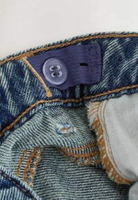 Buză din denim cu un buton din plastic transparent, buclă elastică violet și cusături brune contrastante; margine tocită cu material deschis dedesubt.