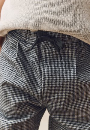 Nahaufnahme einer Person, die schwarz-weiß karierte Hosen mit einem schwarzen Kordelzug trägt, der zu einer Schleife gebunden ist, und einen beige gestrickten Pullover.