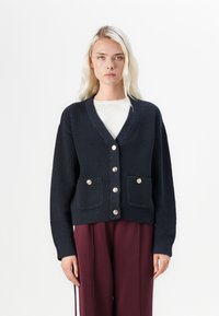 Navy ribbed cardigan with gold buttons, V-neck, and two front pockets. Long sleeves and a textured finish. Paired with maroon trousers.