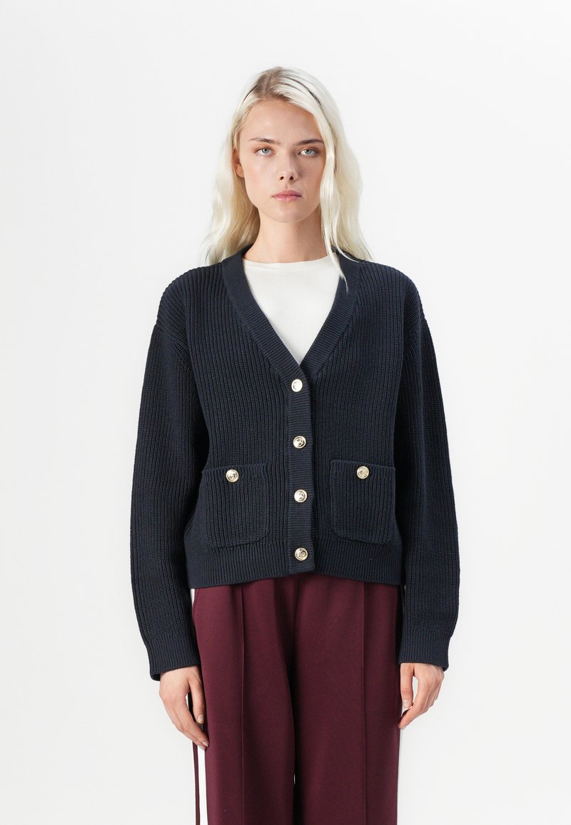Navy ribbed cardigan with gold buttons, V-neck, and two front pockets. Long sleeves and a textured finish. Paired with maroon trousers.