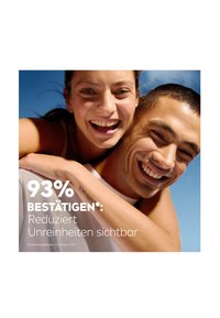 Two people are smiling against a blue background. Text reads "93% BESTÄTIGEN*: Reduziert Unreinheiten sichtbar." They wear casual attire.