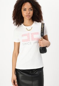 Balta medvilnė t-shirt su rožine ir pilka grafine spauda "EC" ir "SINCE 1998." Derinama su juoda odine sijonu ir siūtu juodu rankine.