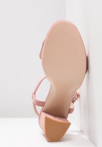 Semelle et talon d'une sandale rose en daim pour femme avec bride à la cheville, présentés contre des murs et un sol blancs.