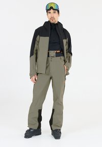 Skiausrüstung mit einer schwarzen und olivgrünen Jacke sowie passenden Hosen. Die Hosen haben einen orangefarbenen Gürtel und schwarze Akzente. Eine Skibrille ist auf dem Kopf getragen.