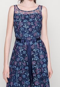 Robe navy avec broderies florales en rose et bleu, col transparent, et une ceinture en satin à la taille. Silhouette en A.