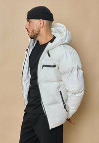 Doudoune blanche avec capuche intégrée, dotée de zips noirs contrastants et d'une texture matelassée, portée sur une chemise noire.