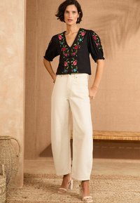 Fekete blúz színes virágos hímzéssel, V-kivágással és puffos ujjal. Krémszínű, bő szárú farmernadrág. Semleges színű, magassarkú szandál. Közelben egy fonott kosár.