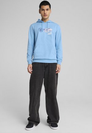 Jasnoniebieska bluza z kapturem z kieszenią na froncie i graficznym nadrukiem, zestawiona z ciemnymi, luźnymi dżinsami i czarnymi sneakersami.