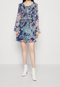 Modrá šaty s paisley vzorem, s volánkovým výstřihem a rukávy, vyrobené z průhledné tkaniny. Nosené s bílými kotníkovými botami.