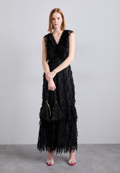 Robe noire avec des détails en franges, décolleté en V, longueur au sol. Modèle tenant un sac à main noir texturé. Chaussures avec des accents et des sangles roses.